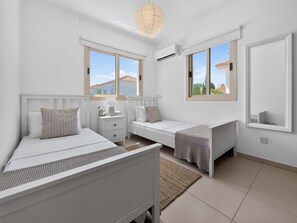 A bedroom with two beds, a bedside table, a lamp, and a window with a view of buildings. - Villa Andreas: Private Pool & Modern Comfort (Protaras)