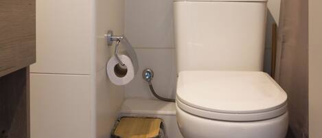 A small, compact bathroom with a toilet, a wooden waste bin, and a roll of toilet paper on the wall.