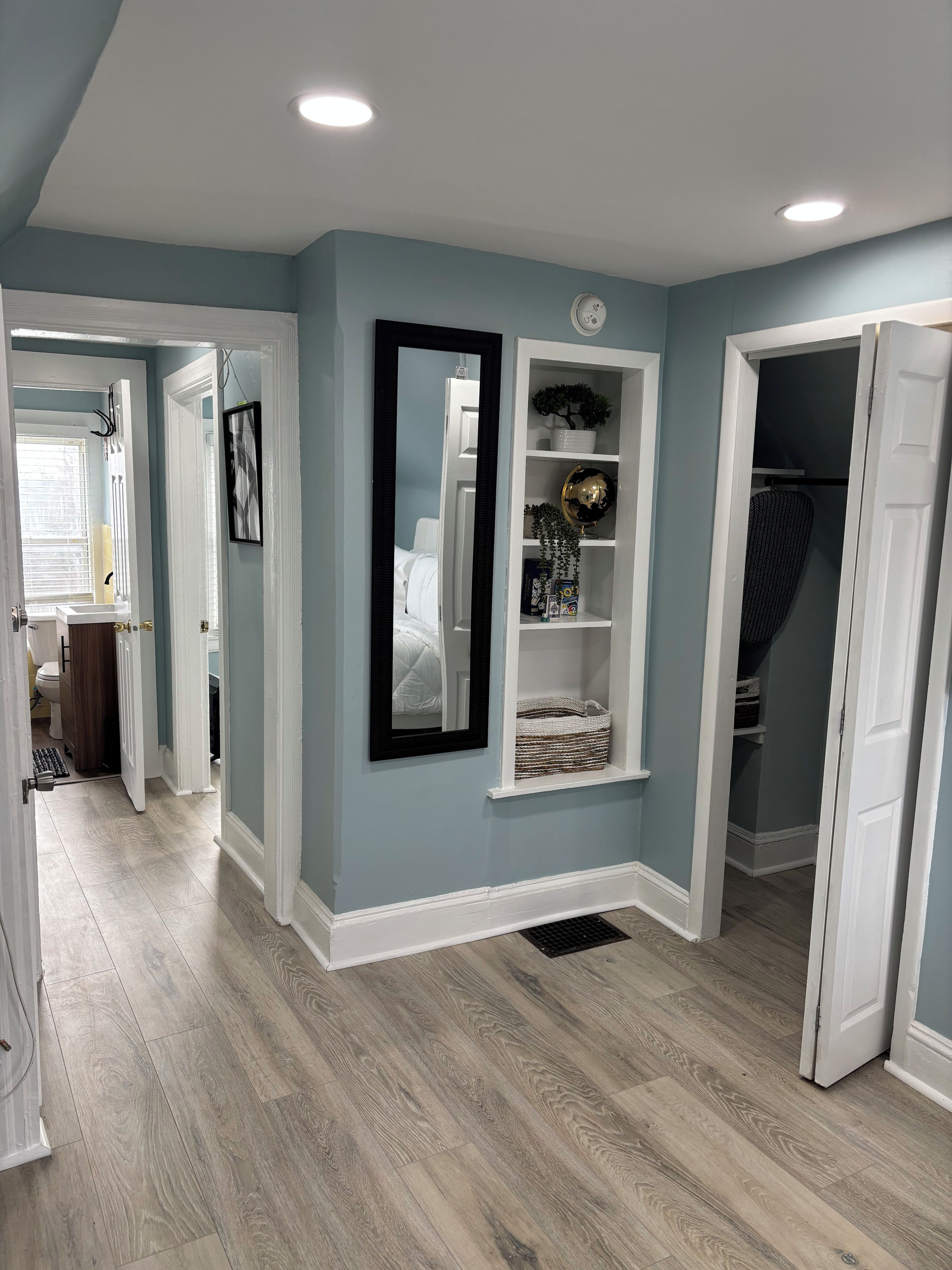 A hallway with a light blue wall, a white door, and a mirror. There is a shelf with decorative items and a globe. The floor is wooden.