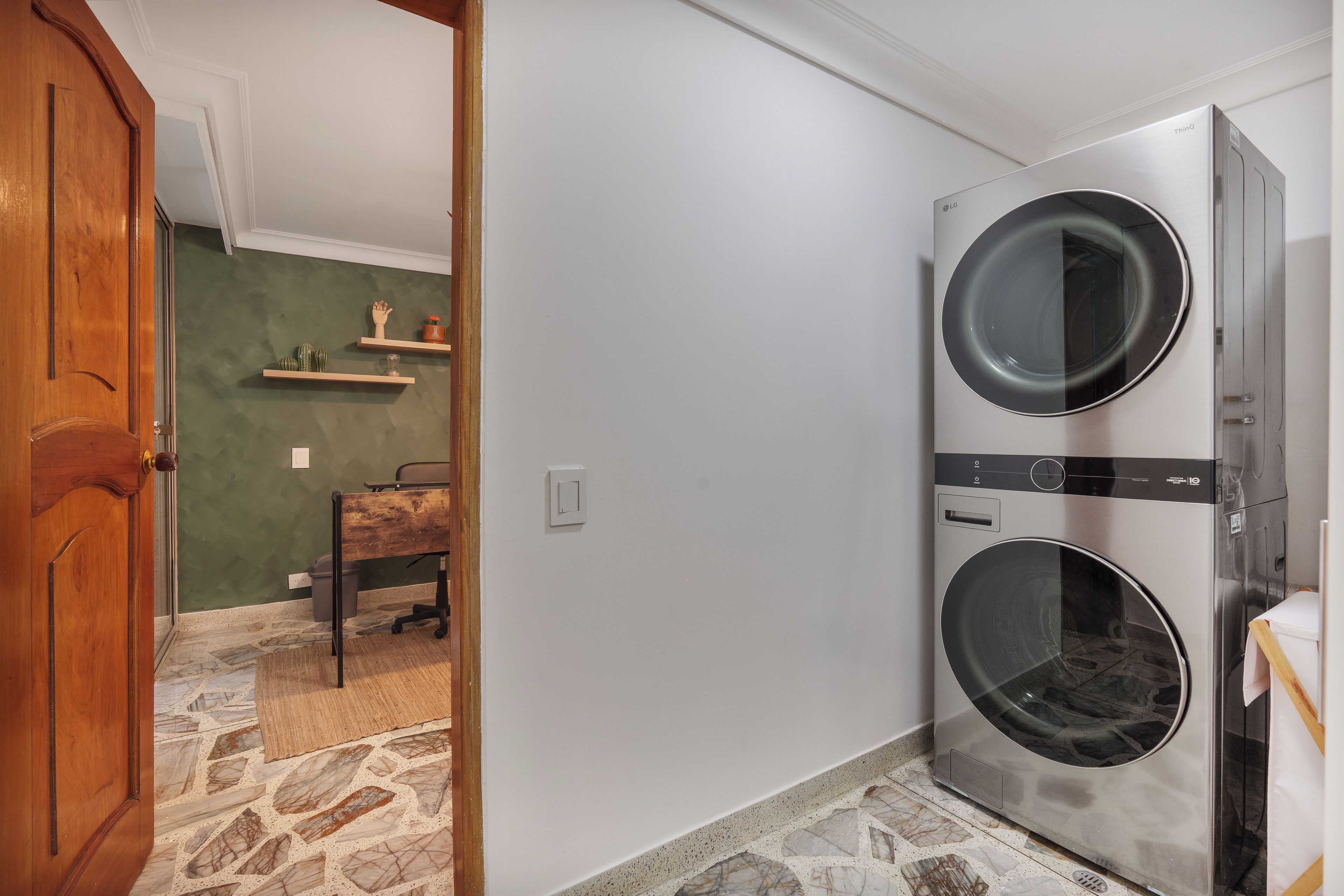 A laundry room with two stacked washing machines, a wooden door, a stone floor, and a small table with a chair.