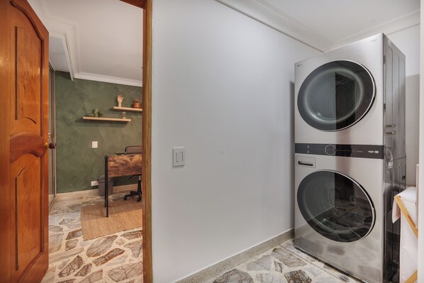 A laundry room with two stacked washing machines, a wooden door, a stone floor, and a small table with a chair. - Peaceful 2-Bedroom Retreat in El Poblado (Medellín)