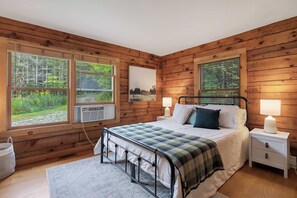 Quarto de cabana de madeira com cama, duas luminárias de cabeceira, mesa de cabeceira e janela com vista para uma floresta.