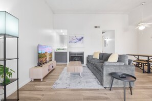 A modern living room with a flat-screen TV, a gray sofa, a coffee table, and a dining area with a wooden table and benches.