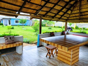 Een overdekt buitengebied met een houten tafel en banken, een rieten dak en uitzicht op een blauw huis en tuin.
