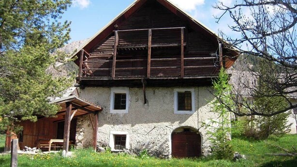 Un edificio rústico de piedra con un balcón de madera, una pequeña estructura adosada de madera y un entorno natural.