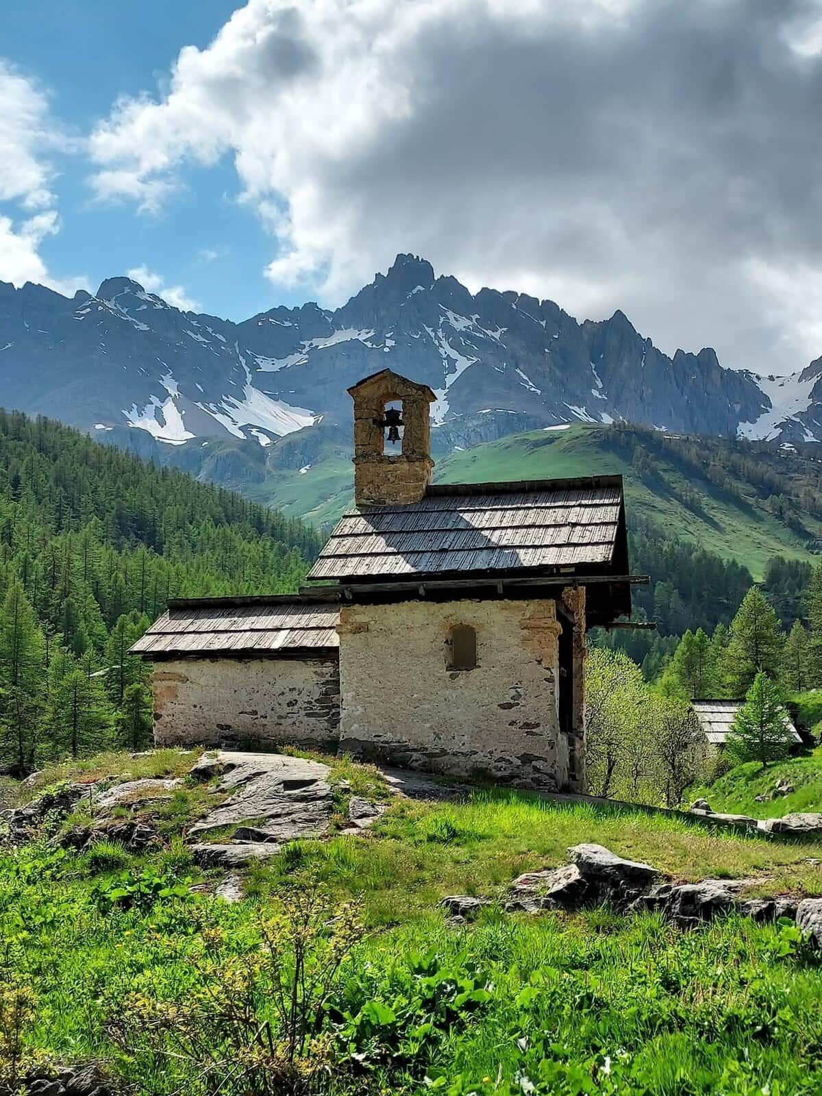 A rustic stone church with a bell tower, surrounded by lush greenery and snow-capped mountains in the background.