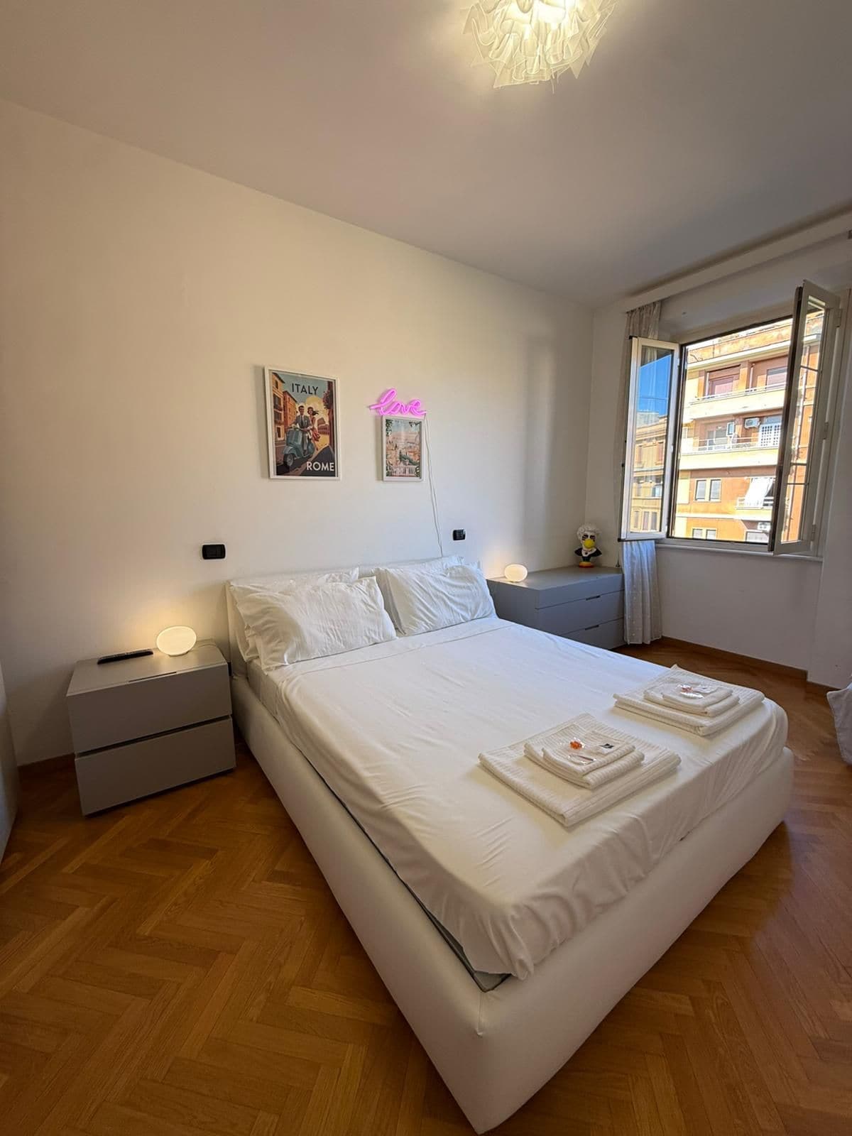 Une chambre d’hôtel comprenant un lit, des tables de chevet, une petite table de nuit avec une lampe, une fenêtre donnant sur des immeubles et deux éléments de décoration murale.