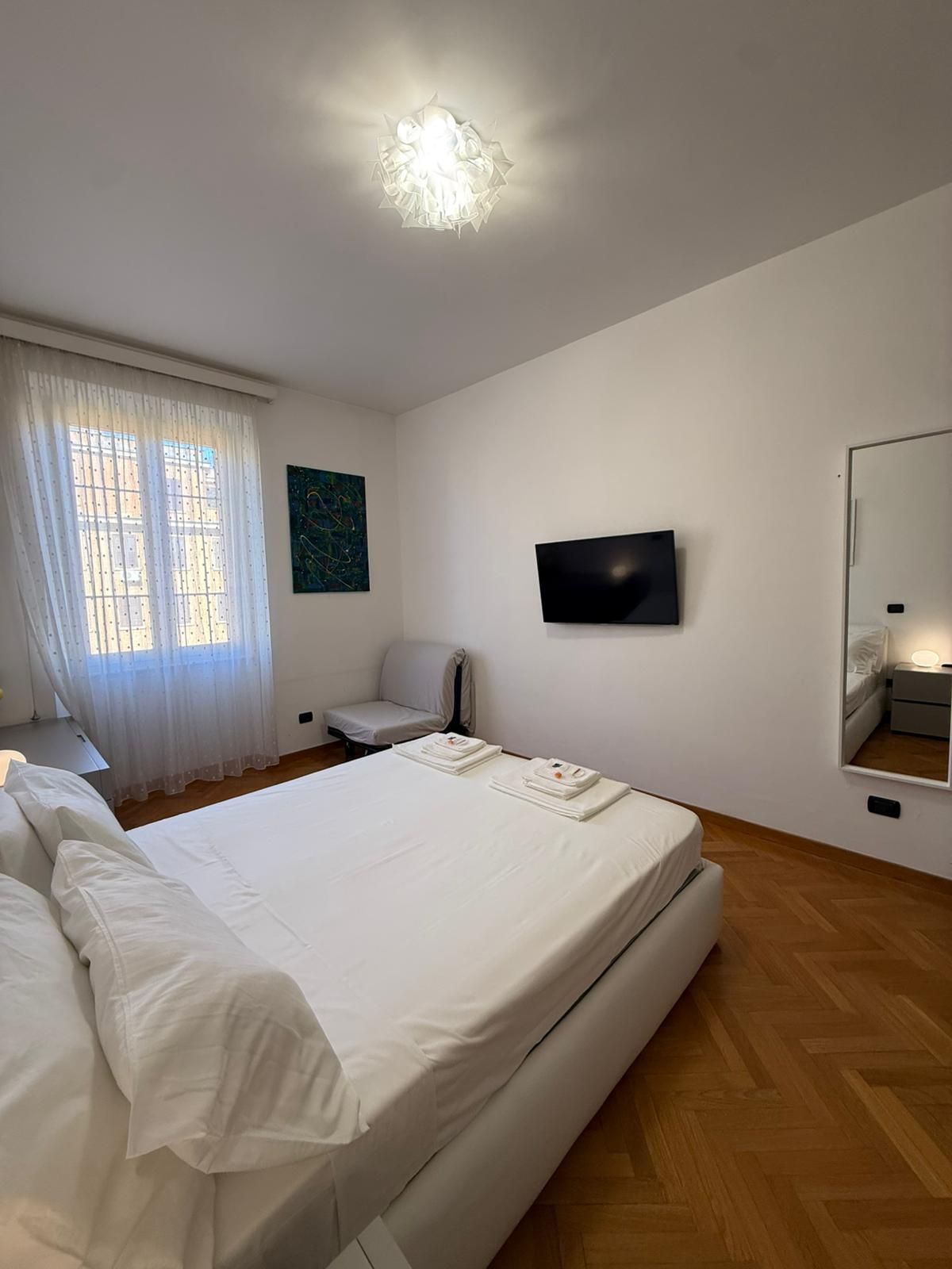 Habitació d'hotel amb un llit, una cadira, un televisor, una finestra amb cortines i un quadre a la paret.