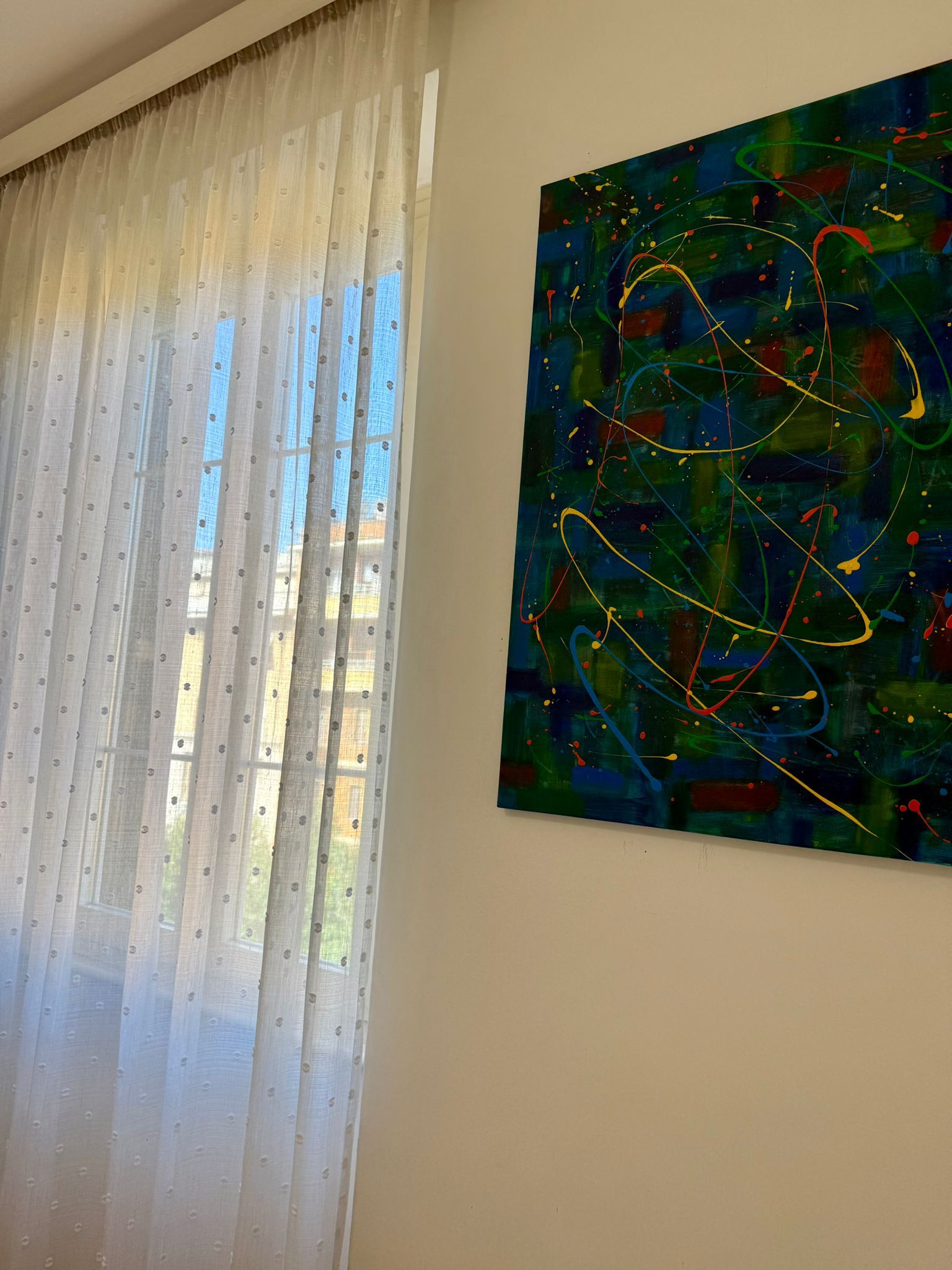 A room with a window covered by sheer curtains and a wall-mounted abstract painting.