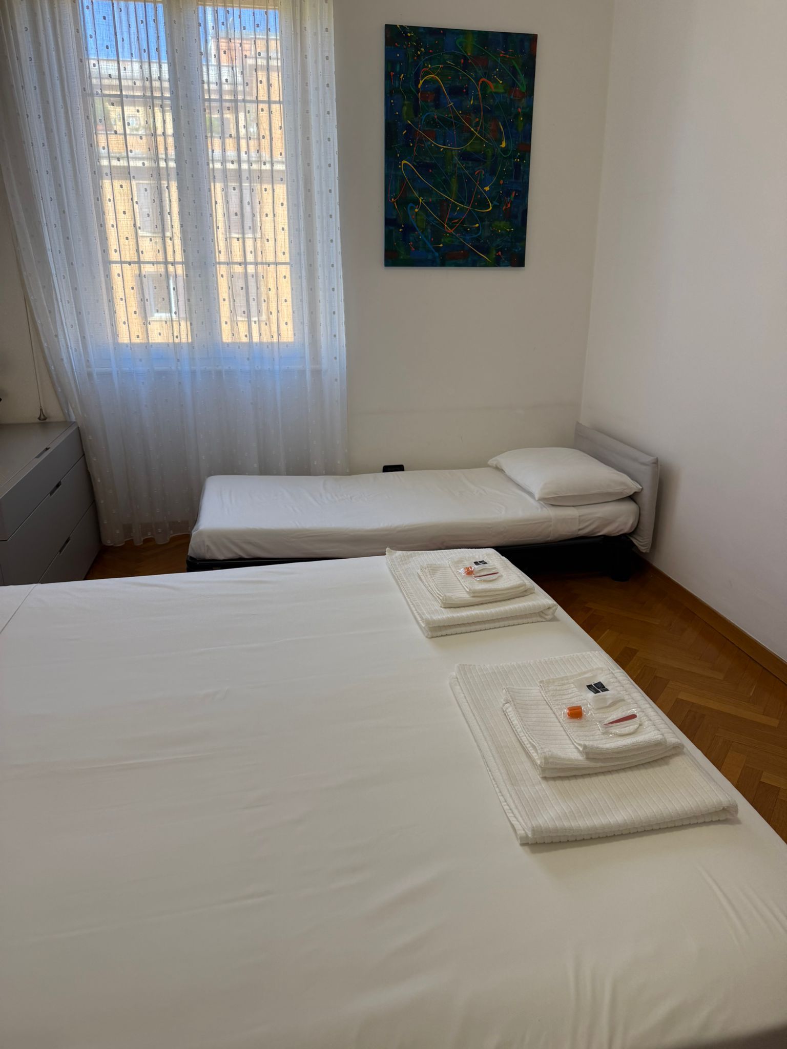 Een hotelkamer met twee bedden, een raam met dunne gordijnen, een schilderij aan de muur en een houten vloer.