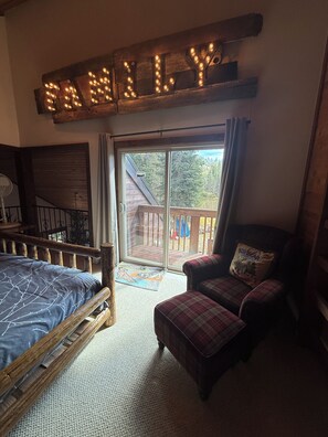 A bedroom with a bed, a chair, a couch, a wooden sign above the bed, and a sliding glass door leading to an outdoor deck. - Charming 3-bedroom cabin in delightful Soda Springs getaway. Sorry no pets. (Soda Springs)