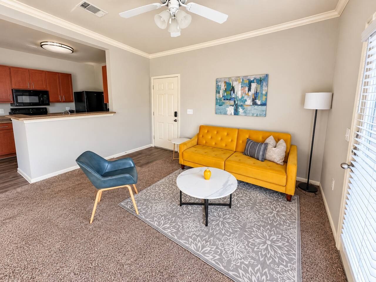 A living room with a yellow sofa, a blue chair, a white coffee table, and a kitchen area in the background.