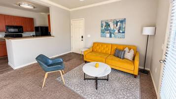 A living room with a yellow sofa, a blue chair, a white coffee table, and a kitchen area in the background.