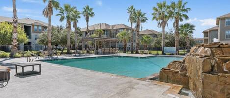 A swimming pool with a stone waterfall feature, surrounded by palm trees and lounge chairs.
