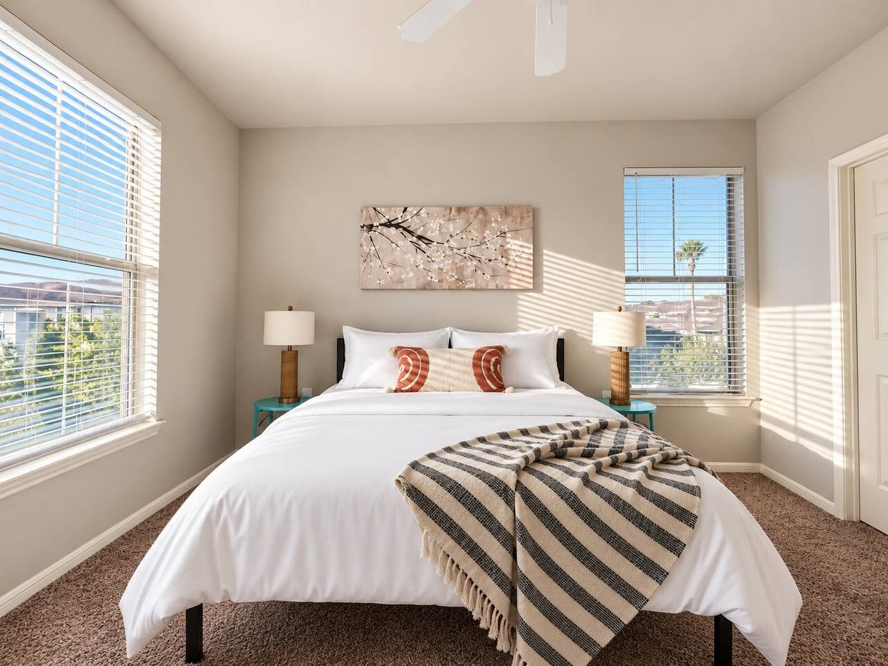 A bedroom with a large bed, two bedside tables with lamps, a window with blinds, and a wall-mounted artwork.