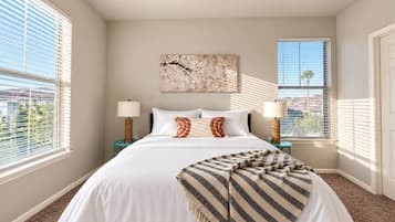 A bedroom with a large bed, two bedside tables with lamps, a window with blinds, and a wall-mounted artwork.