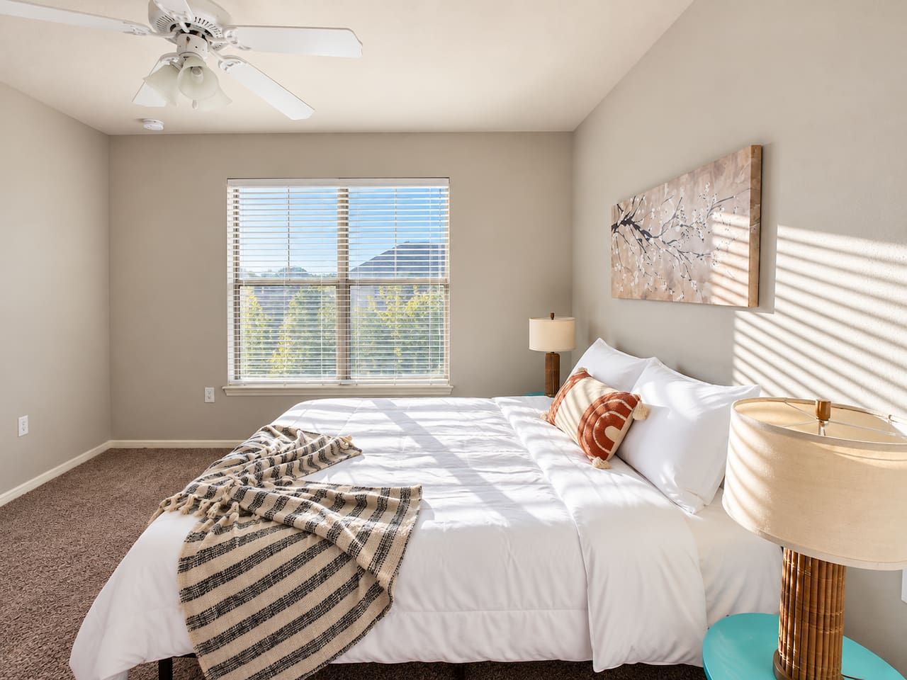 A bedroom with a bed, a ceiling fan, a window with blinds, a lamp, and a wall-mounted artwork.