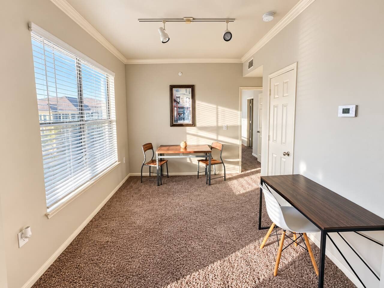 A room with a dining table, chairs, a window with blinds, a framed picture on the wall, and a thermostat.