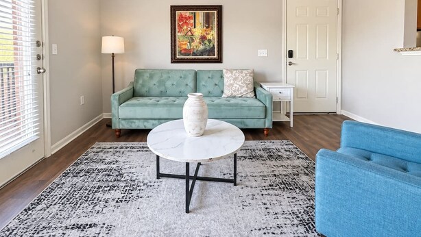 Una sala de estar con un sofá verde, una mesa blanca con un florero, un tapete estampado y un cuadro en la pared.