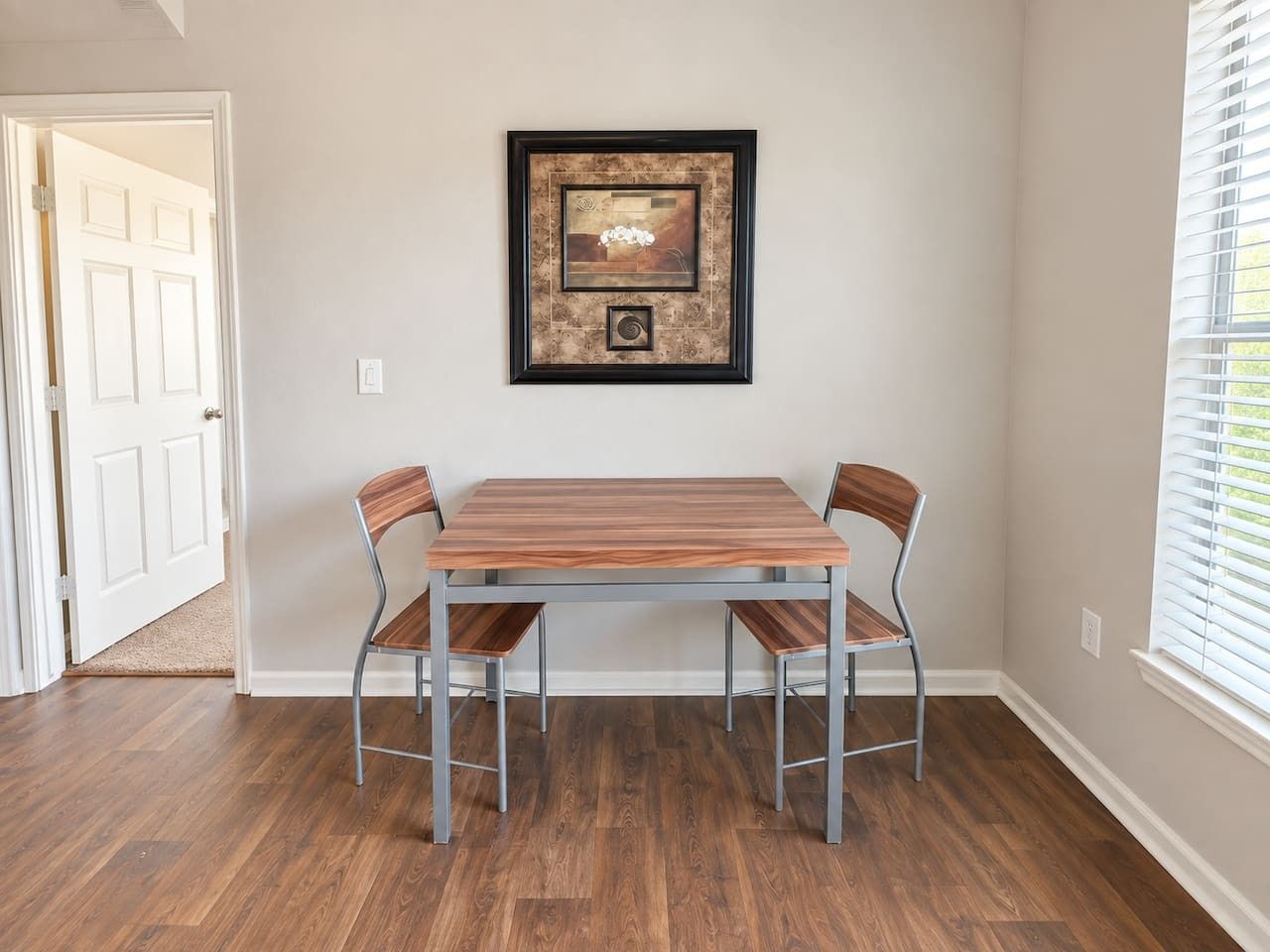 A dining area with a table and chairs, a framed artwork on the wall, and a window with blinds.