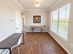 A room with a dining table and chairs, a framed picture on the wall, and a window with blinds. - Sleepover | Modern 1BD/1BA + Gym - Corpus Christi (Corpus Christi)
