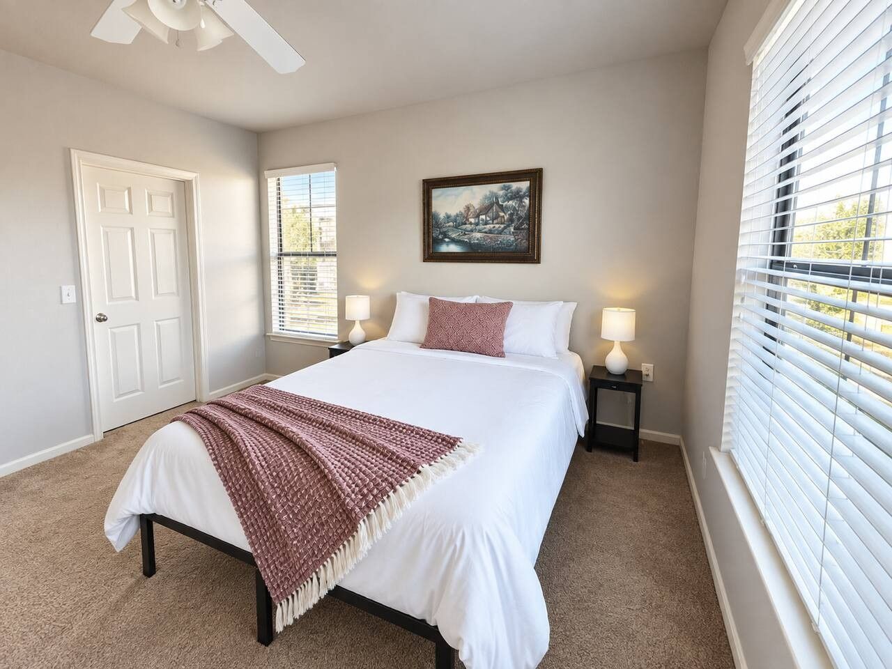 A bedroom with a bed, bedside lamps, a door, a window with blinds, and a painting on the wall.