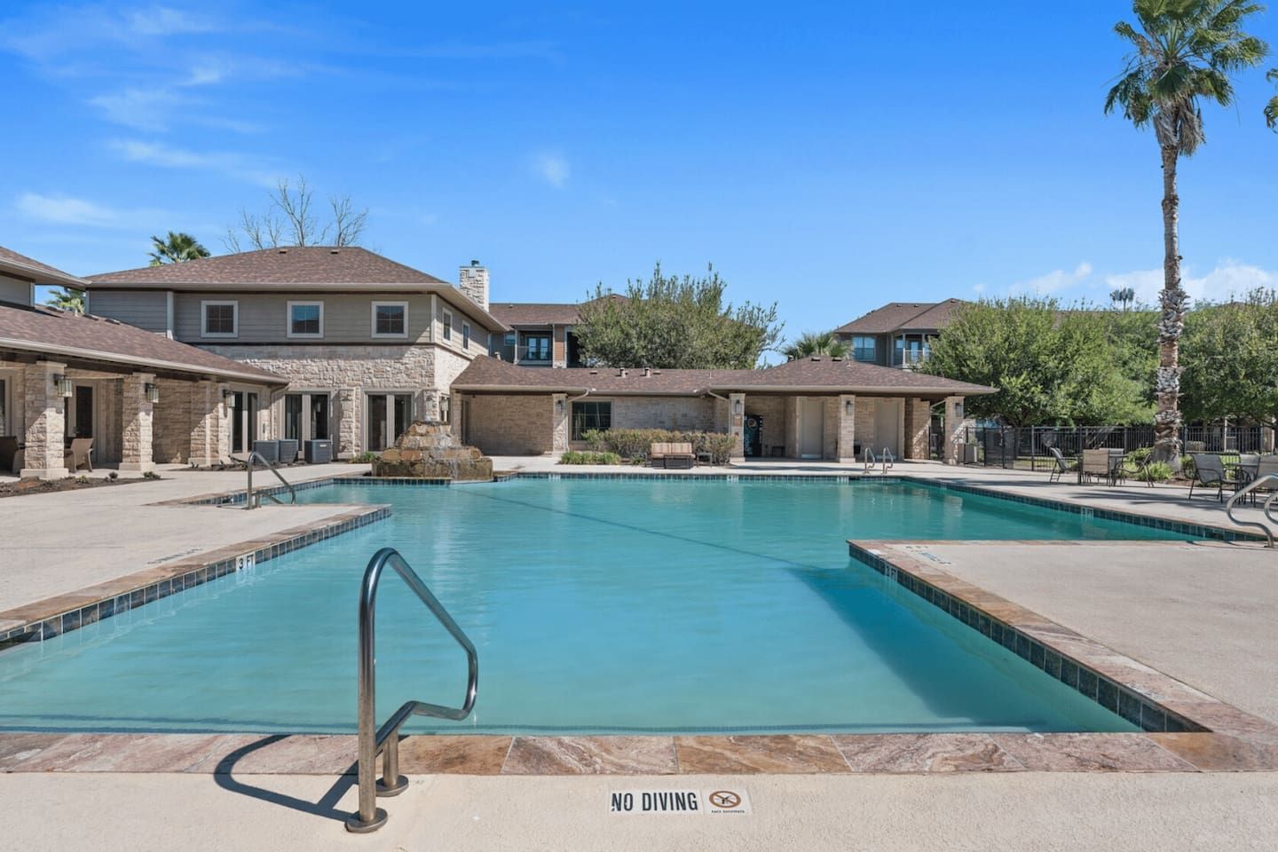 A swimming pool with a no diving sign, surrounded by houses with stone facades and a clear blue sky.