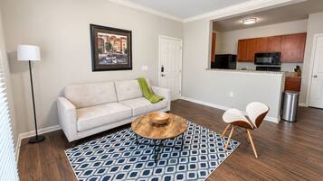 A living room with a sofa, coffee table, and chairs, a patterned rug, and a kitchen in the background.