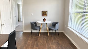 A room with a round table, two chairs, a door, a window with blinds, and a framed picture on the wall.