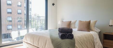 Une chambre avec un grand lit, une vue sur les bâtiments environnants et une fenêtre équipée de stores.