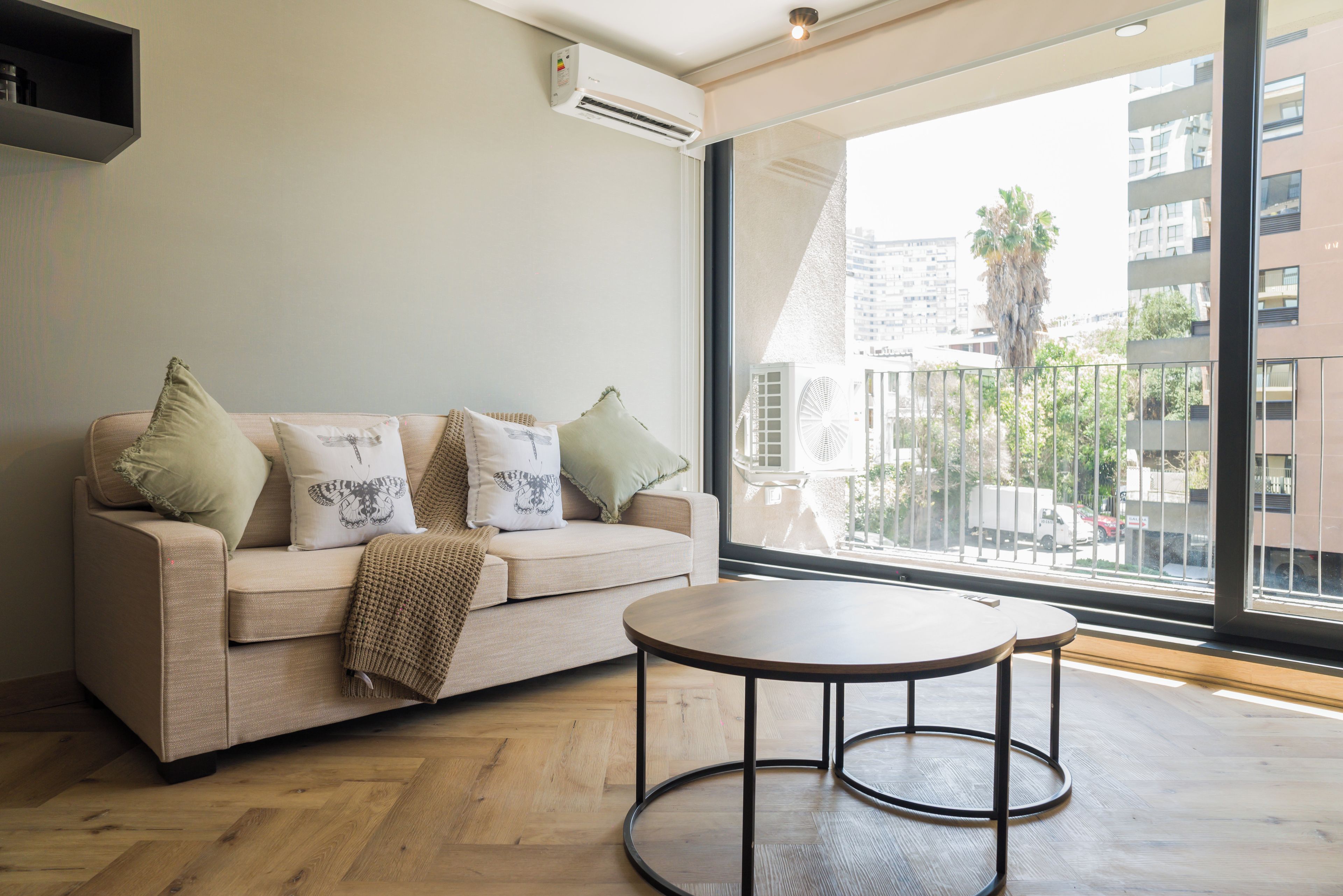 A modern living room with a sofa, pillows, a round table, and a large window with a view of buildings and a palm tree.