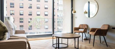Uma sala de estar moderna com um sofá, duas poltronas, uma mesa redonda e uma janela grande com vista para um edifício.