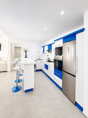 A modern kitchen with white and blue cabinetry, a stainless steel refrigerator, and a bar area with stools. - Panoramic Atlantic Ocean view, first-row location | 2 minutes from the beach (Agadir)