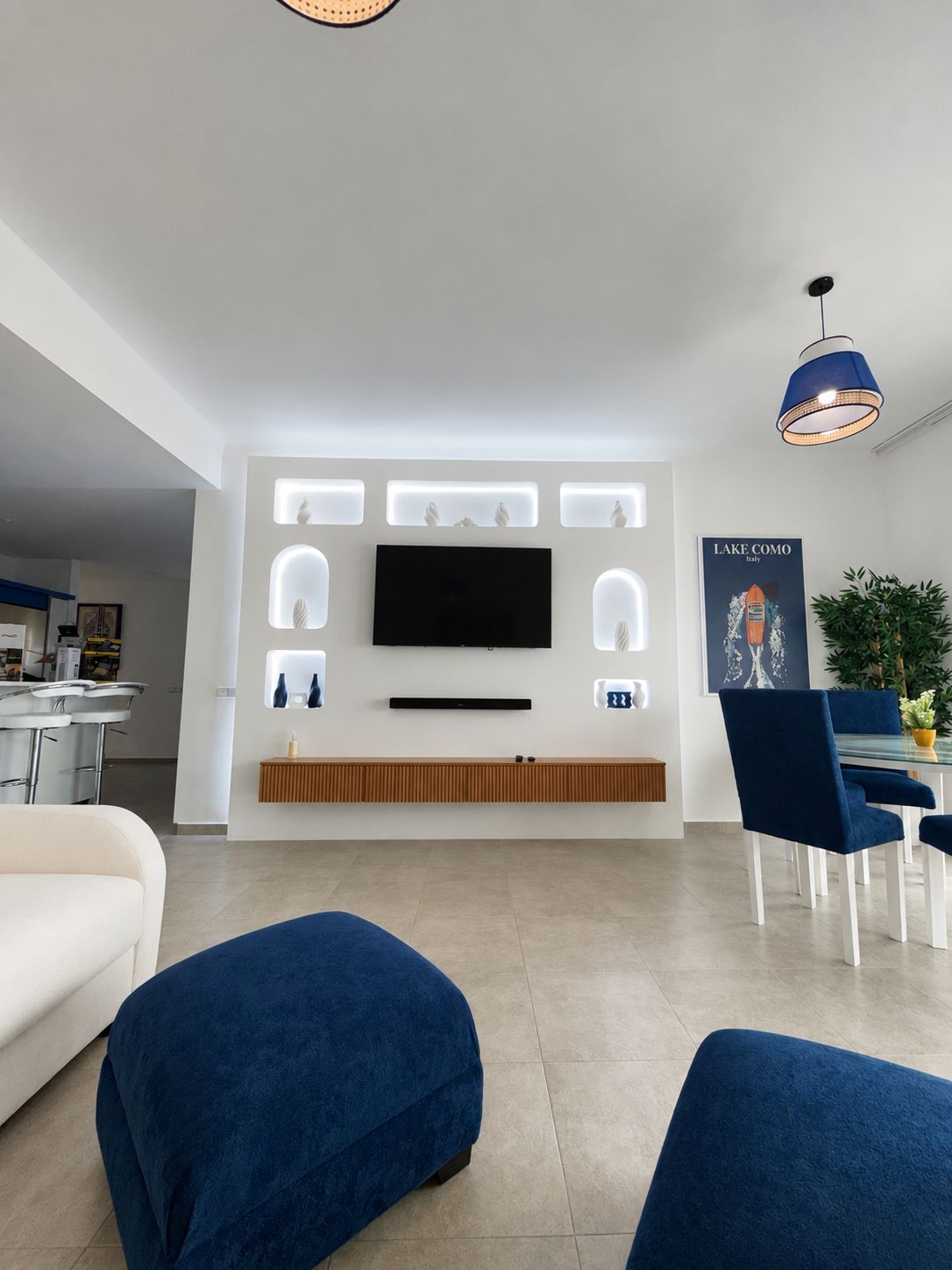 Una sala moderna con un televisor de pantalla plana montado en la pared, un comedor con sillas azules y una cocina al fondo.