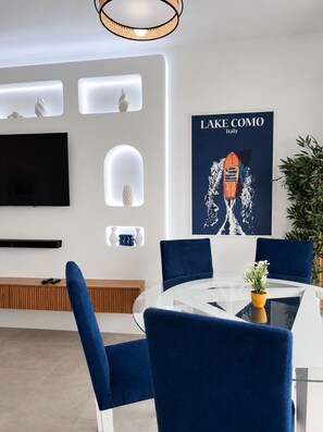 A modern dining area with a round glass table, blue upholstered chairs, a mounted flat-screen TV, and a poster of a boat on Lake Como. - Panoramic Atlantic Ocean view, first-row location | 2 minutes from the beach (Agadir)