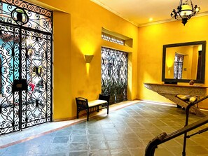 Un couloir avec un mur jaune, un portail en métal, un banc, un miroir et un escalier.