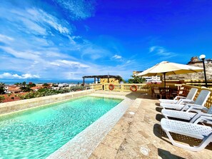 Une piscine sur le toit, avec une eau d’un bleu limpide, entourée de chaises longues et de parasols, offrant une vue panoramique sur le paysage urbain côtier.