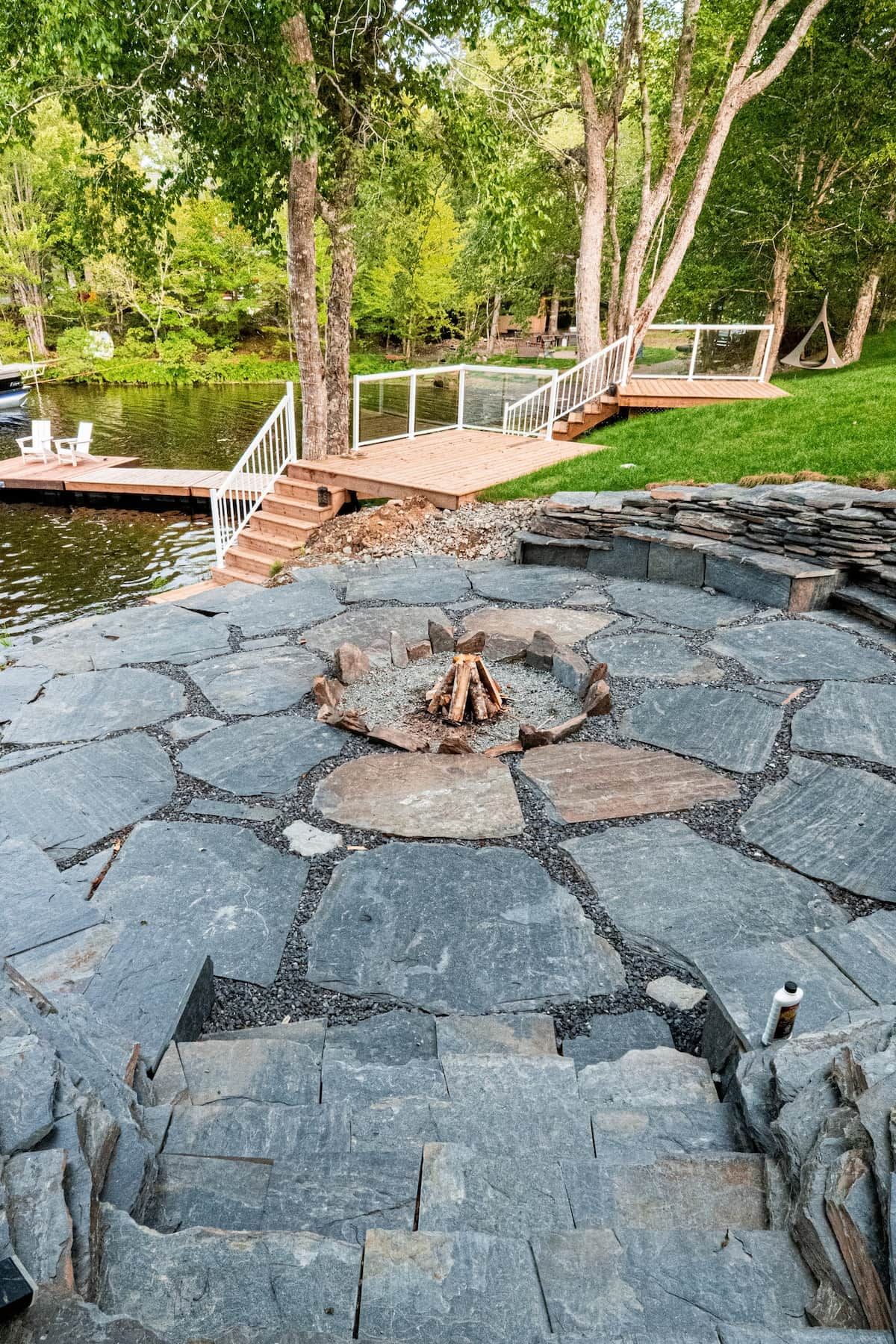 A stone deck with a fire pit in the center, surrounded by a stone wall, and a wooden deck with a railing overlooking a body of water.