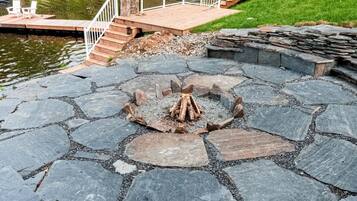 A stone deck with a fire pit in the center, surrounded by a stone wall, and a wooden deck with a railing overlooking a body of water.
