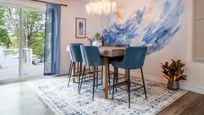 Un espace repas comprenant une table, des chaises, un lustre, une grande peinture abstraite et une vue sur l’extérieur, visible à travers une porte vitrée.