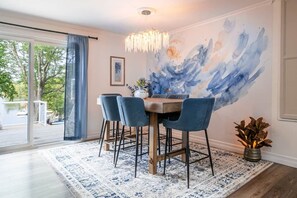 Uma área de refeições com mesa, cadeiras, um lustre, uma grande pintura abstrata e vista através de uma porta de vidro.