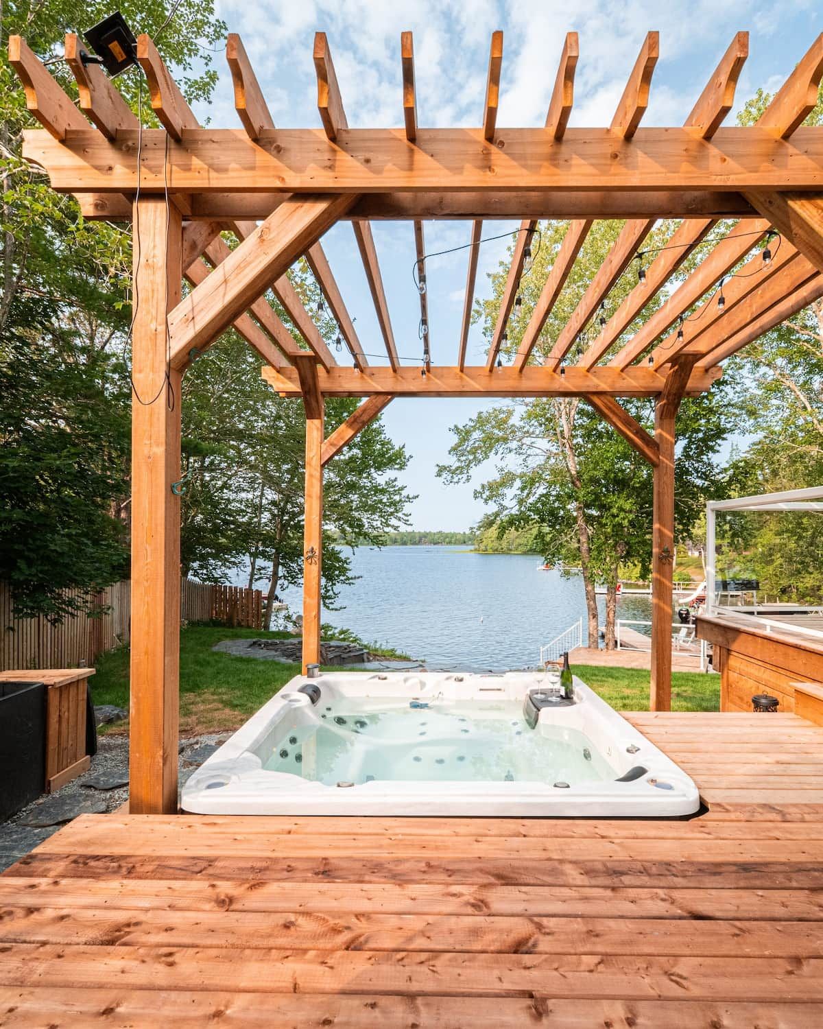 A wooden deck with a hot tub and a pergola overlooking a lake.