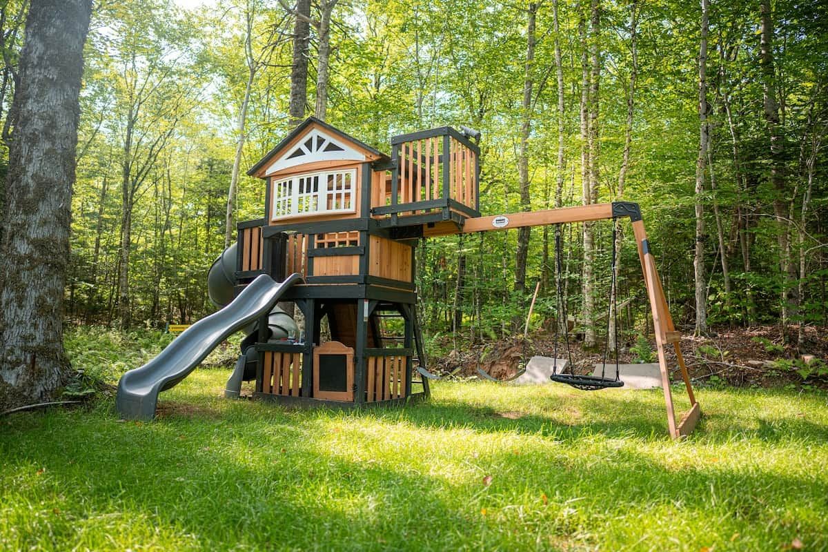 A wooden playhouse with a slide and swing set in a forested area.