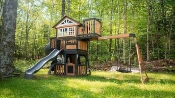 A wooden playhouse with a slide and swing set in a forested area.