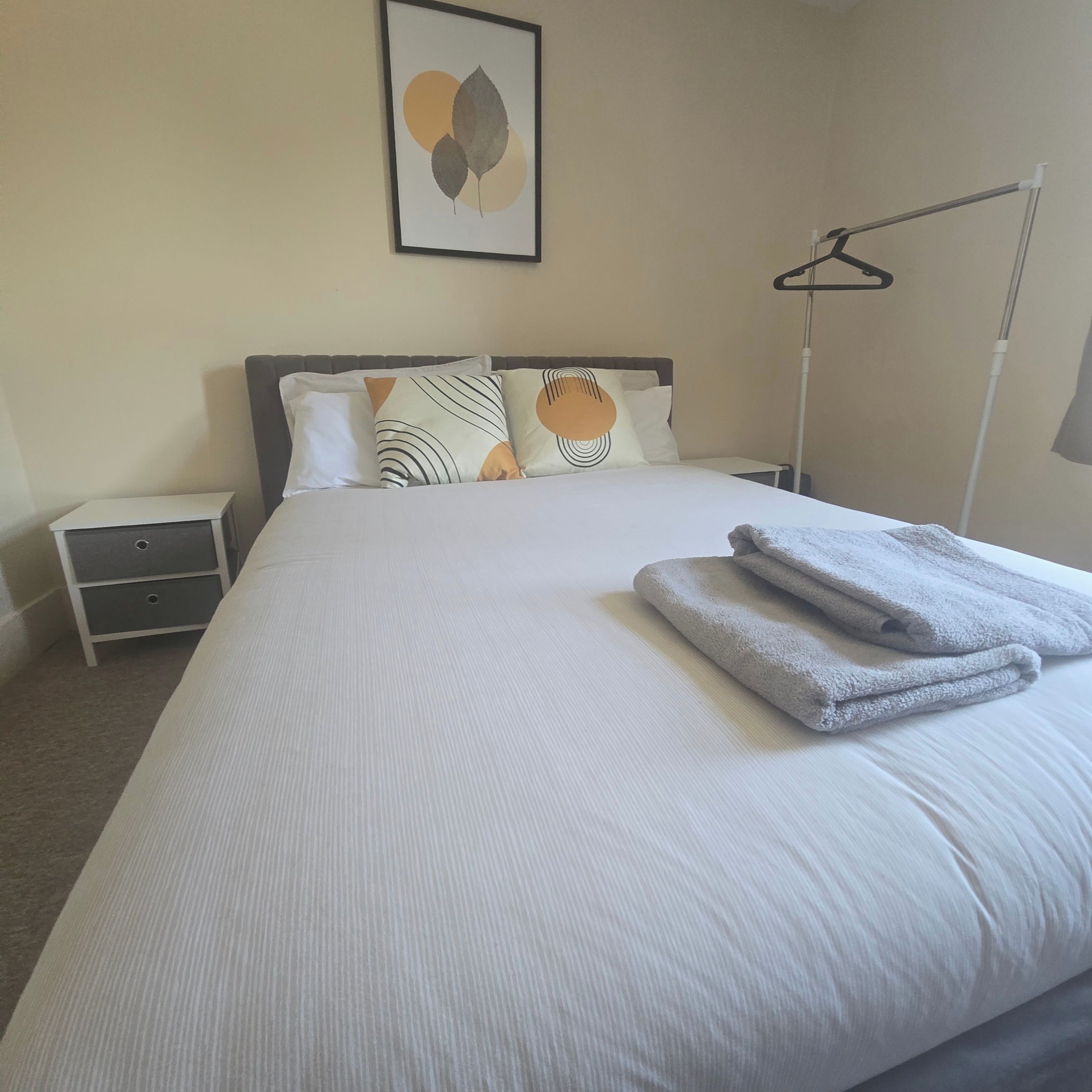 A neatly made bed with a folded towel, a bedside table with a lamp, and a framed artwork on the wall.