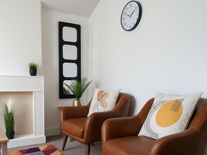 Een woonkamer met twee bruine leren fauteuils, een zwarte wandklok en een witte schoorsteenmantel met een plant.