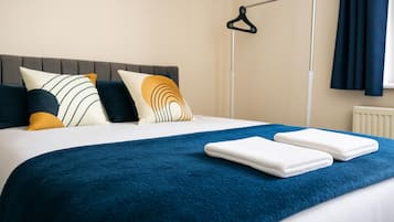 Una cama bien hecha con una manta azul y dos toallas dobladas, un cabecero gris y un perchero negro en la pared.