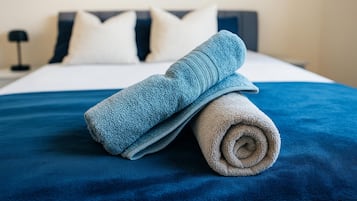 Een net opgemaakt bed met een blauw-witte sprei, twee kussens en een opgerolde handdoek.