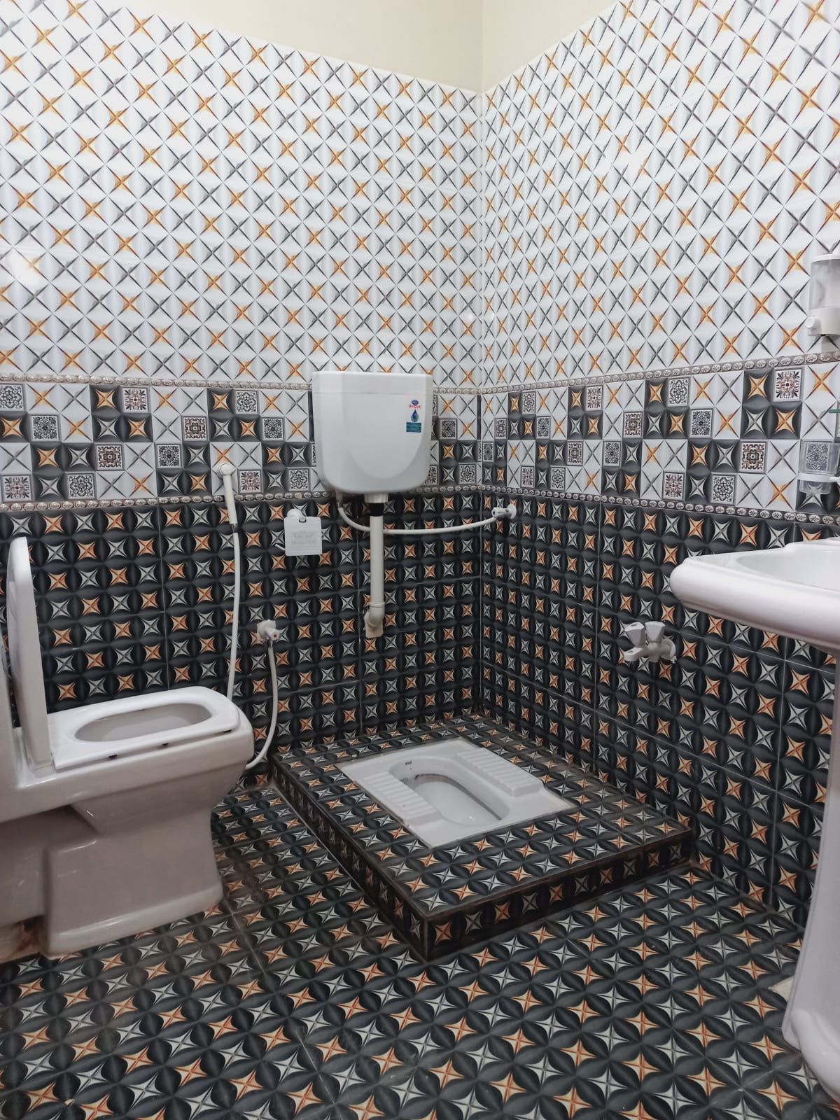 A small bathroom with a toilet, bidet, and sink. The walls are tiled with a geometric pattern.