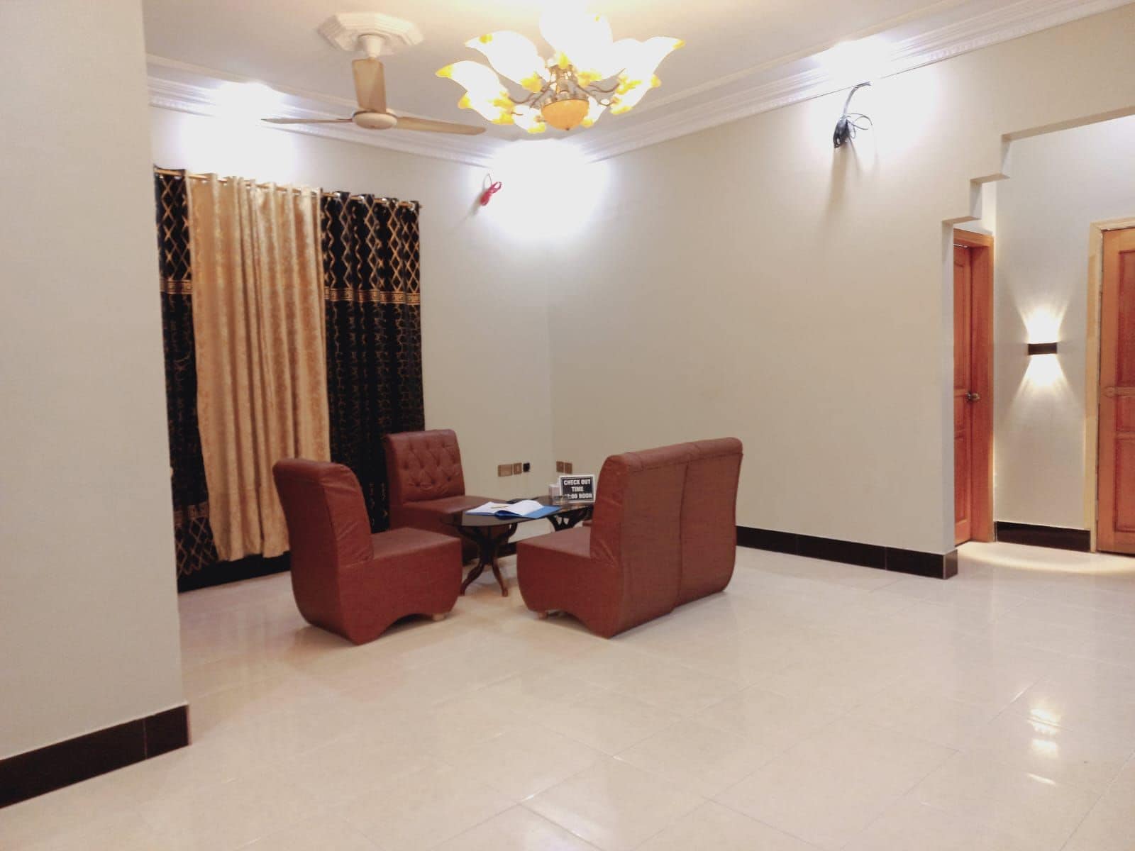 A spacious living room with a high ceiling, a ceiling fan, and a chandelier. There are two red armchairs and a small table with a laptop. The room has a large window with patterned curtains.